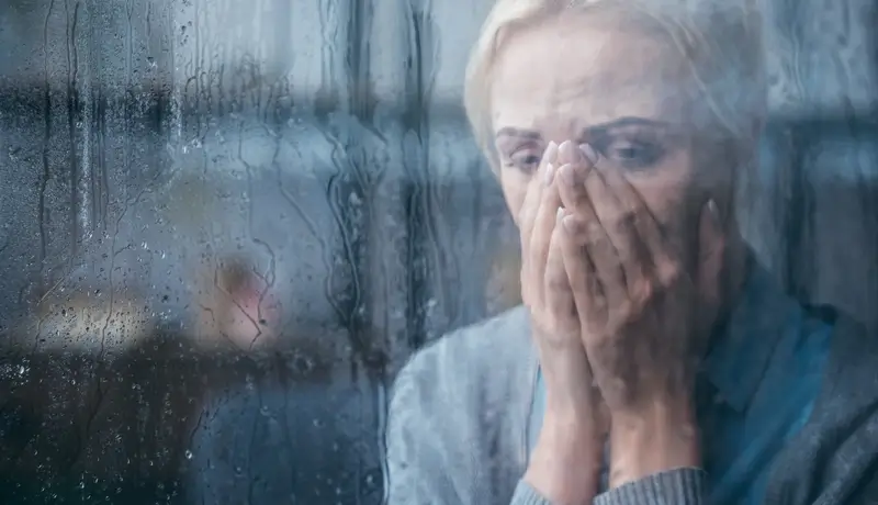 A distressed woman standing behind a rain-covered window, holding her hands to her face in deep emotional pain.