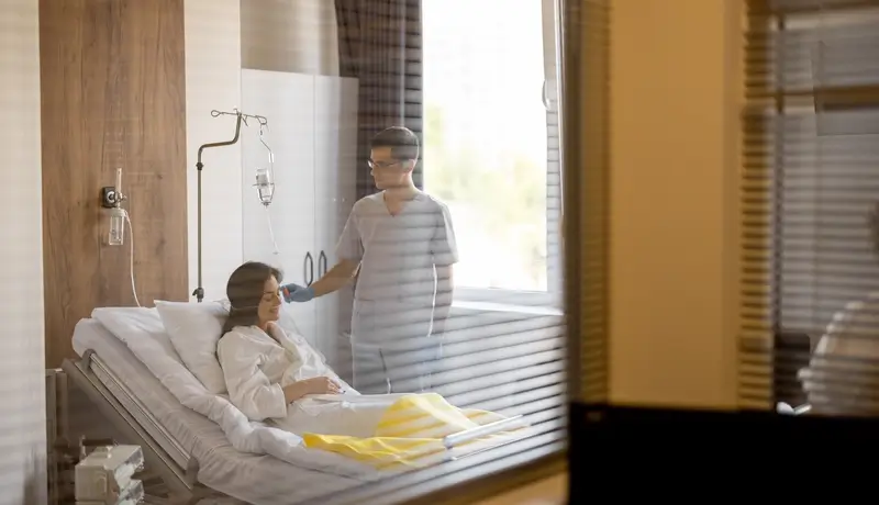 A private hospital room with a patient in a white gown standing near a window, showcasing a clean and organized medical setting.