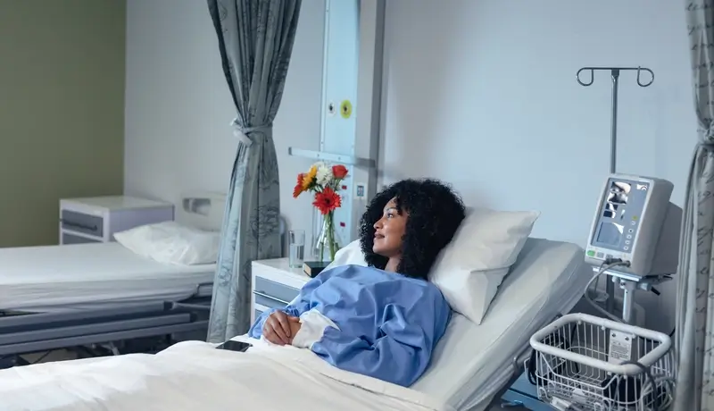 Patient in a hospital bed looking out, surrounded by a calm environment with flowers and medical equipment.