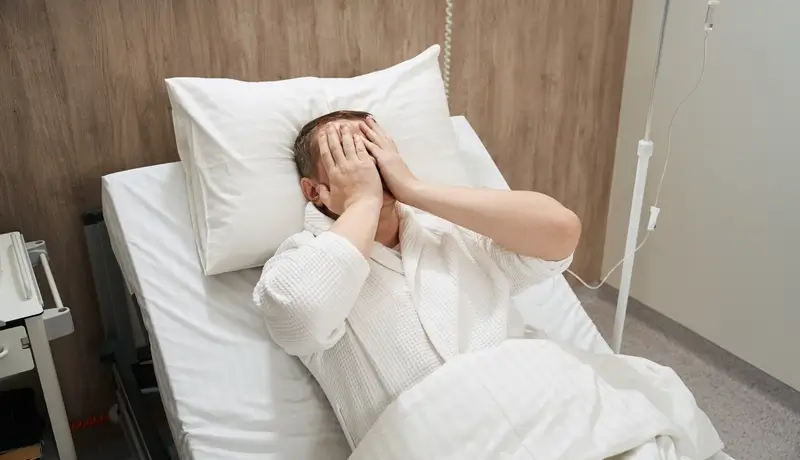 Patient lying in a hospital bed covering their face, appearing stressed or uncomfortable.