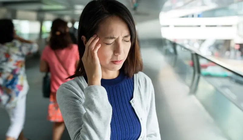 Woman in a public place holding her head, indicating discomfort from a headache.